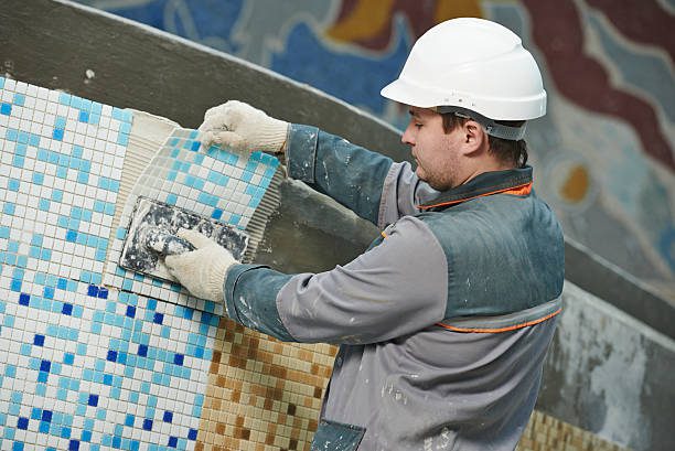 Worker in a hard hat installing blue and white mosaic tiles on a wall using adhesive.